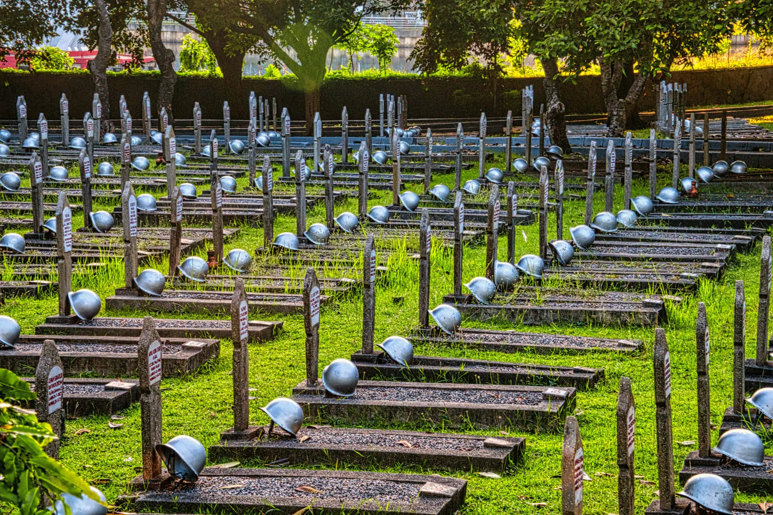 Tombstones with military hardhats located on grassy ground near tall green tall trees in national main heroes cemetery in Indonesia