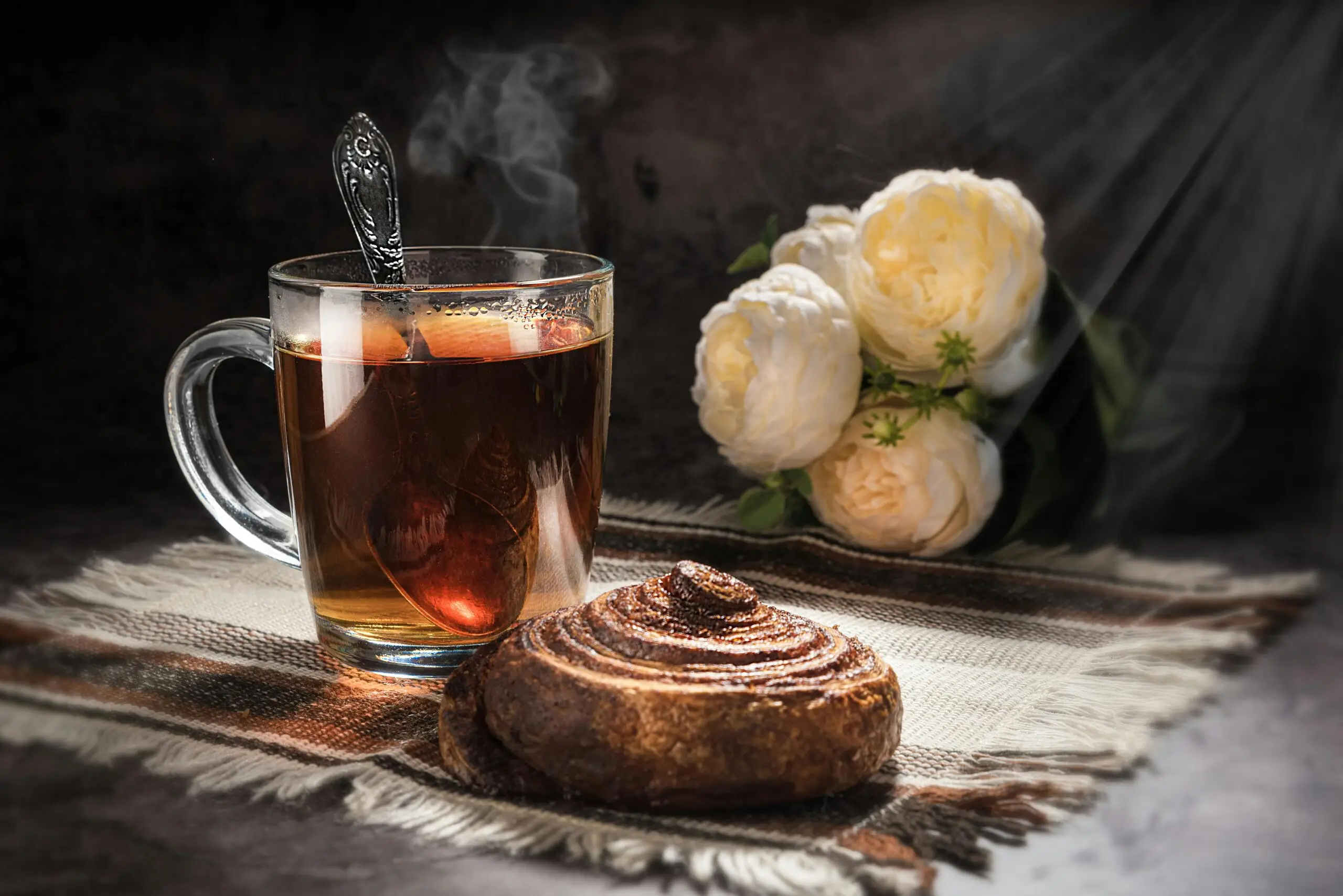 A steaming cup of tea and a cinnamon roll on a table, with white roses in soft lighting.