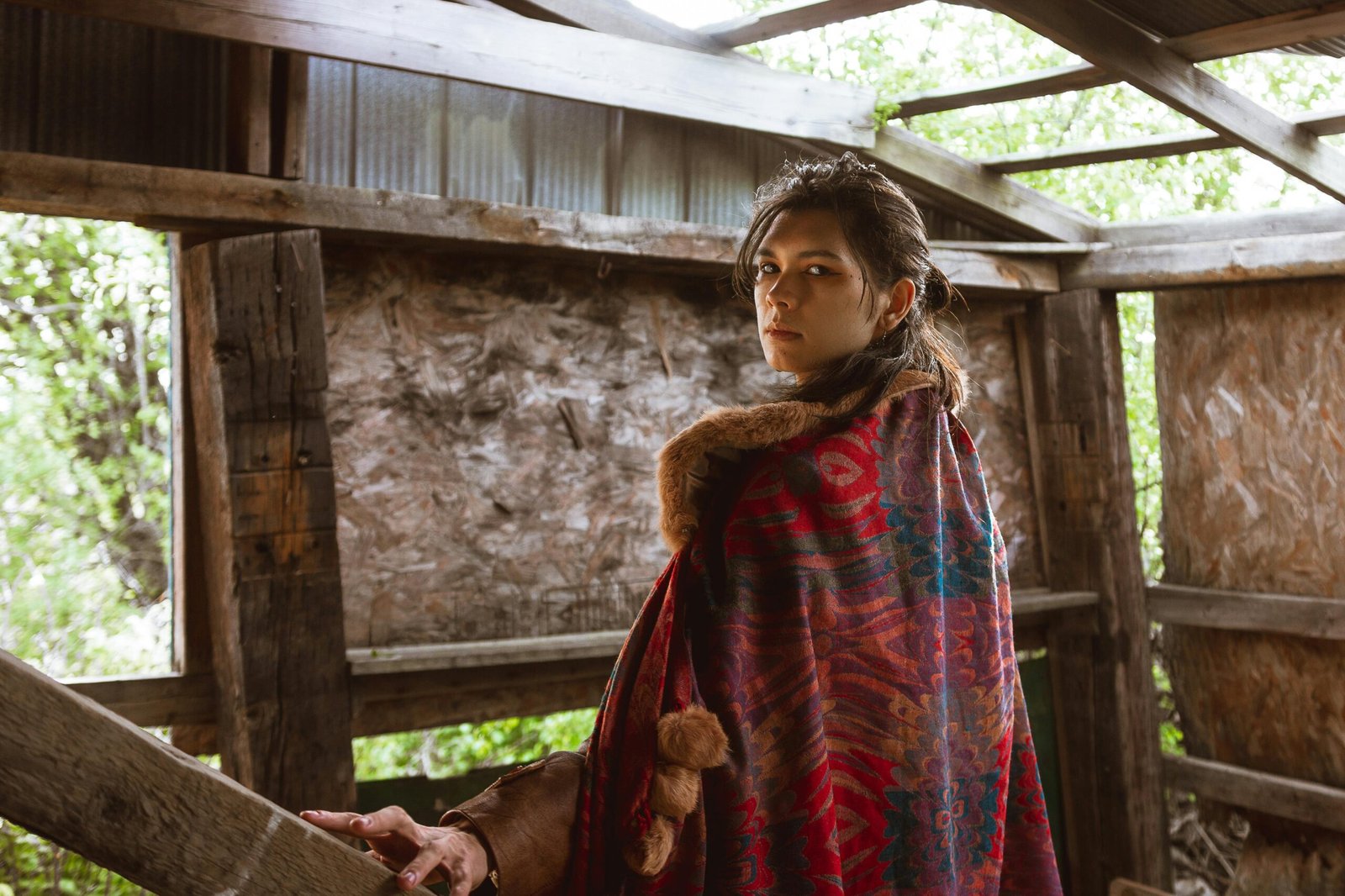 A woman in a colorful cloak stands in a rustic shed, partially covered by greenery.