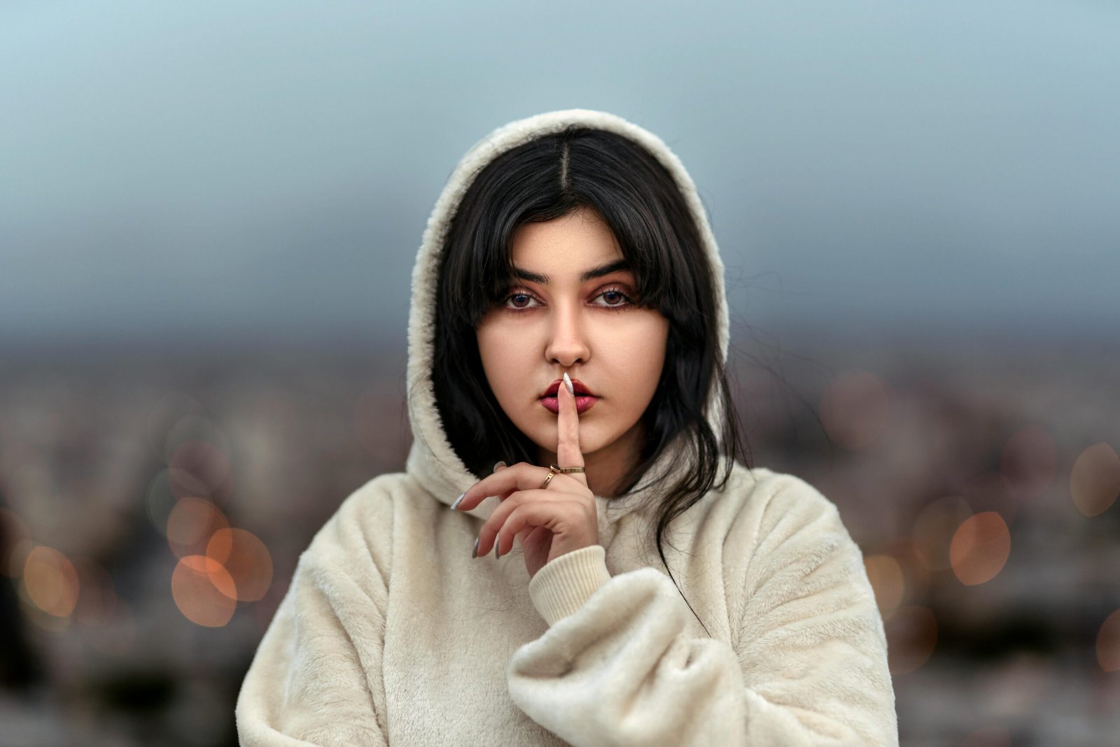 Middle Eastern woman in a hoodie signals silence, standing outdoors with an evening backdrop.