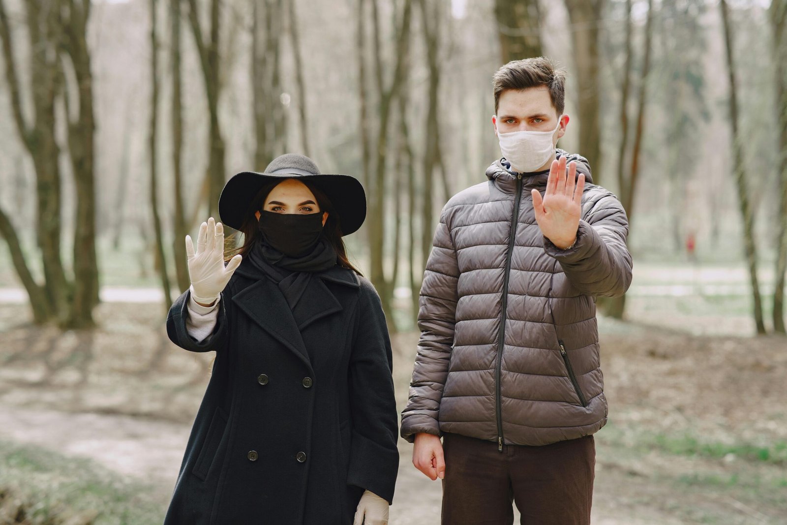 Serious couple in outerwear standing in park in medical masks and latex gloves and showing stop virus gesture while looking at camera