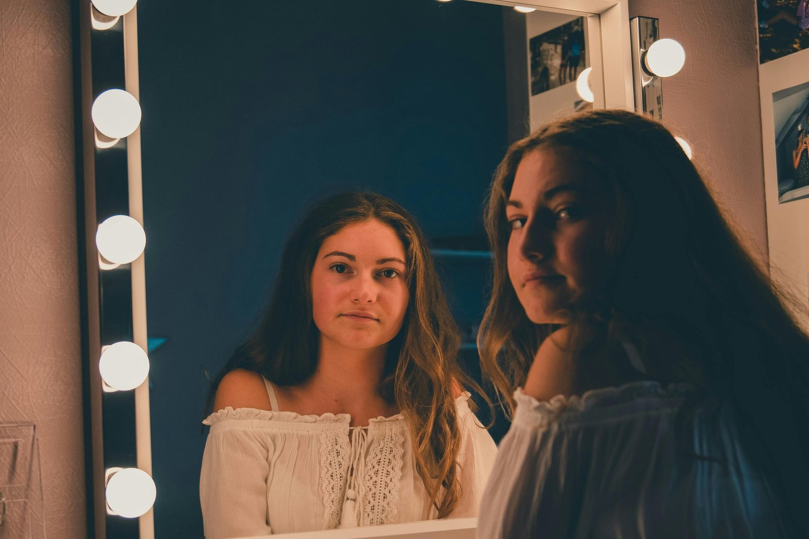 A young woman gazes thoughtfully into a vanity mirror in a softly lit room.