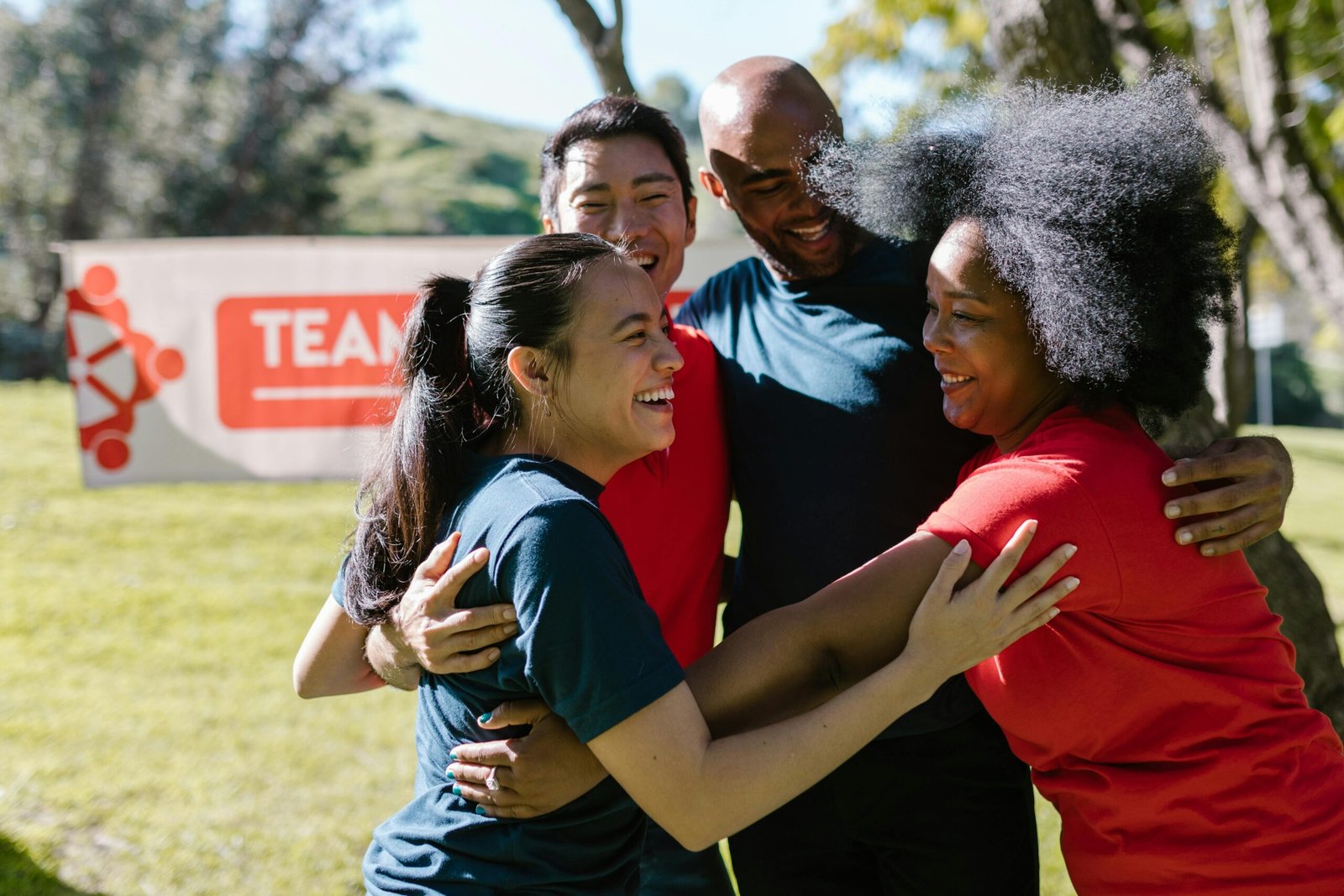 A joyful, diverse group embraces during an outdoor team building event in a sunny park.