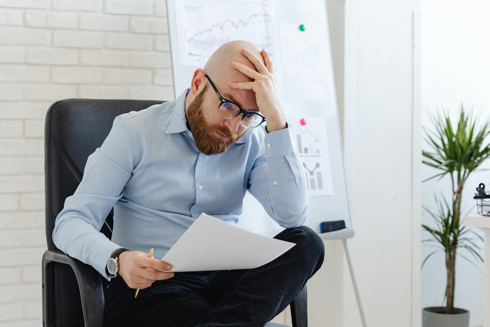 Bald man with glasses sitting in office looking worried at document.